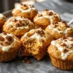 A close-up of two freshly baked pumpkin cream cheese muffins with golden-brown tops and swirls of cream cheese filling visible on the surface, displayed on a rustic wooden plate with a few cinnamon sticks and a sprig of mint for garnish. The muffins are soft and moist, with a slight crackled crust and a hint of pumpkin flavor.
