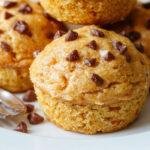 A close-up of three golden-brown pumpkin cream cheese muffins on a rustic white plate, topped with a swirl of cream cheese filling that glistens slightly. The muffins have a moist, tender crumb with a slight crack on top revealing the creamy filling inside. Fresh orange pumpkin slices and a sprinkle of cinnamon are arranged around the plate, enhancing the autumnal setting. Soft natural light highlights the warm tones and inviting texture of the muffins.