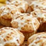 A close-up of golden-brown banana cream cheese muffins arranged on a rustic wooden platter. The muffins have a slightly cracked top revealing a creamy filling inside, with a light dusting of powdered sugar. Fresh banana slices and a drizzle of cream cheese glaze are visible as garnishes, creating an inviting and appetizing presentation.