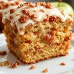 A slice of Apple Spice Cake topped with a generous swirl of brown sugar icing, showcasing moist, spiced apple cake layers with visible chunks of apple and a glossy, caramel-colored icing on top, set on a rustic wooden platter with a light dusting of cinnamon around the edges.