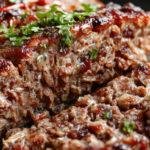 A sliced Korean BBQ meatloaf sits on a white plate, glazed with a glossy sweet and spicy sauce. The meatloaf has a golden-brown crust with visible bits of green scallions and sesame seeds on top. Juices glisten around the slices, showing a tender, moist interior, plated with a side of steamed vegetables and rice in the background.