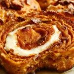 A sliced loaf of swirled pumpkin cream cheese bread sits on a rustic wooden table, revealing its moist, swirled interior with streaks of cream cheese and vibrant orange pumpkin. The top crust is golden-brown with a slight sheen, and a generous slice is lifted to showcase the tender crumb and marbled swirl inside.