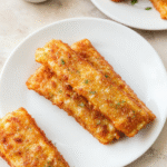 Golden, crispy mozzarella sticks with bubbly breading, plated on a white plate with a small bowl of marinara, sprinkled with parsley. The platter rests on a warm wooden table, softly lit, with the gooey cheese peeking from the crust and a hint of parmesan.