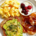 A vibrant breakfast plate featuring fluffy scrambled eggs folded with cottage cheese, a small bowl of Greek yogurt with berries, oats prepared with milk, a slice of avocado on toasted whole grain bread, and a side of turkey bacon, sprinkled with green herbs on a light wood table.